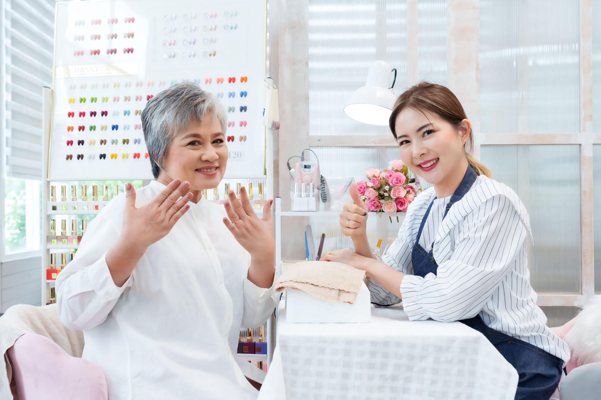 Senior Asian woman happily displaying freshly manicured hands next to young Asian nail technician inside cheerful small nail spa salon decorated with bright nail polish displays and flowers